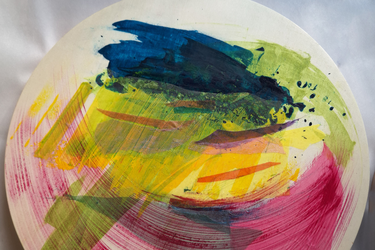 A round abstract art print or decorative plate featuring colorful collage and brushstrokes in blue, green, yellow, pink, and orange on a white background, photographed against a light gray fabric backdrop.